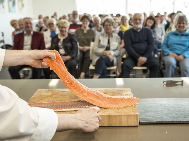 Fischkochstudio Koch präsentiert frisches Lachsfilet vor interessiertem Publikum in moderner Showküche.
