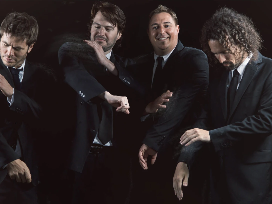 twistem_16_9.jpg Vier Männer in dunklen Anzügen und Krawatten, die lächelnd und selbstbewusst Staub abklopfen.