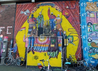 Mural Das Bild zeigt ein farbenfrohes Wandgemälde in Köln mit stilisierten Figuren, die Kleidung in leuchtenden Farben tragen. Fahrräder sind vor der gemalten Wand geparkt.