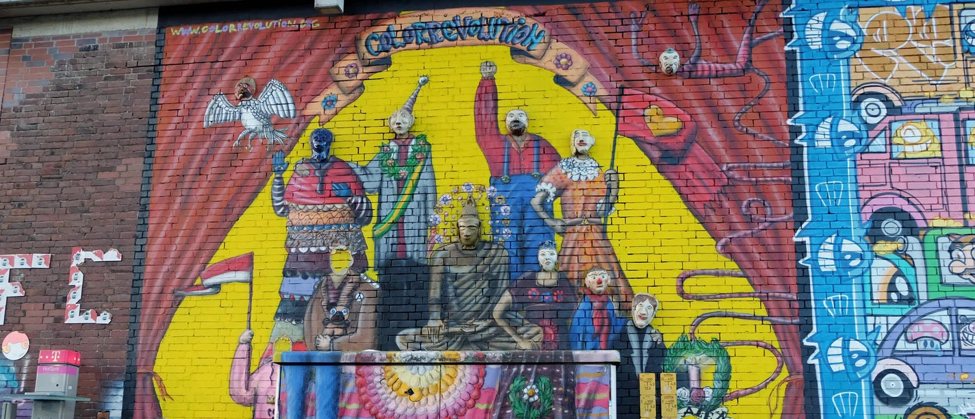 Mural Das Bild zeigt ein farbenfrohes Wandgemälde in Köln mit stilisierten Figuren, die Kleidung in leuchtenden Farben tragen. Fahrräder sind vor der gemalten Wand geparkt.