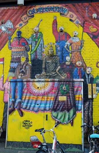 Mural Das Bild zeigt ein farbenfrohes Wandgemälde in Köln mit stilisierten Figuren, die Kleidung in leuchtenden Farben tragen. Fahrräder sind vor der gemalten Wand geparkt.The picture shows a colourful mural in Cologne with stylized figures wearing clothes in bright colors. Bicycles are parked in front of the painted wall.
