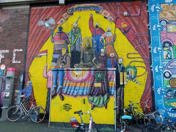 Mural Das Bild zeigt ein farbenfrohes Wandgemälde in Köln mit stilisierten Figuren, die Kleidung in leuchtenden Farben tragen. Fahrräder sind vor der gemalten Wand geparkt.