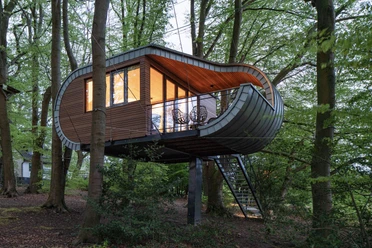 Ein modernes Baumhaus mit Holzverkleidung und Panoramafenstern, umgeben von dichtem Wald.