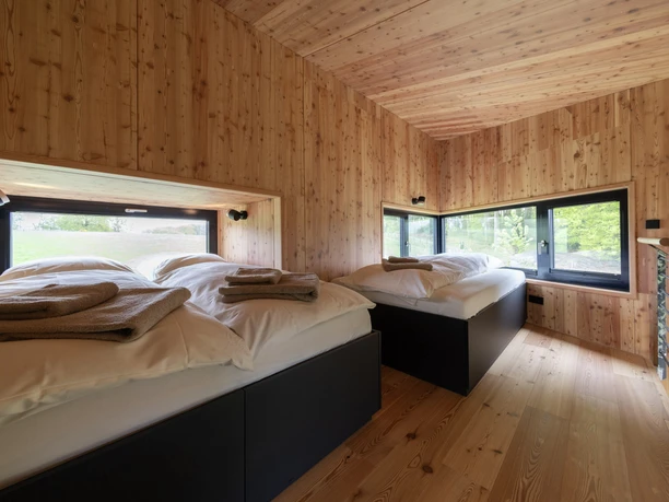Innenansicht Baumhaus Zwei moderne Einzelbetten in einem lichtdurchfluteten, hölzernen Raum mit großen Fenstern und Naturblick.