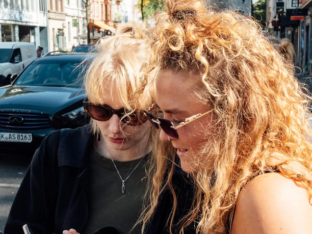 Mindwalk in Köln Zwei junge Frauen mit Sonnenbrillen stehen auf einer belebten Straße und schauen auf ein Mobiltelefon.