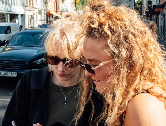 Mindwalk in Köln Zwei junge Frauen mit Sonnenbrillen stehen auf einer belebten Straße und schauen auf ein Mobiltelefon.Two young women wearing sunglasses are standing on a busy street looking at a cell phone.