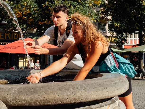 Mindwalk in Köln Zwei Personen schöpfen an einem historischen Brunnen Wasser. Die Hände sind ausgestreckt.