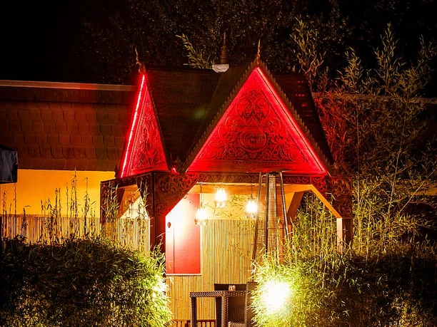 RoyalThai (13).jpg Rotes thailändisches Pavillon bei Nacht, umgeben von Bambus und beleuchtetem Weg, wirkt einladend.