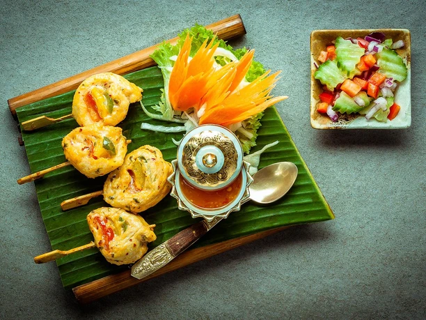 RoyalThai (1).jpg Drei Spieße mit frittierten Happen auf Bananenblatt, dekoriert mit geschnitztem Gemüse und Dip-Sauce.