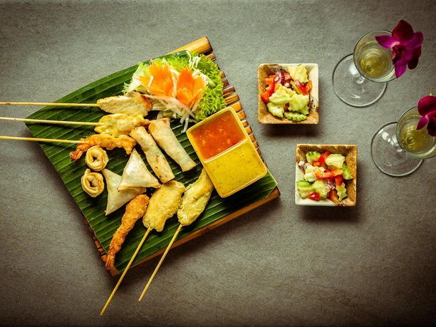 RoyalThai (7).jpg Thailändische Vorspeisenplatte auf Bananenblatt, umgeben von Salat, Soßen und dekorativen Orchideen.