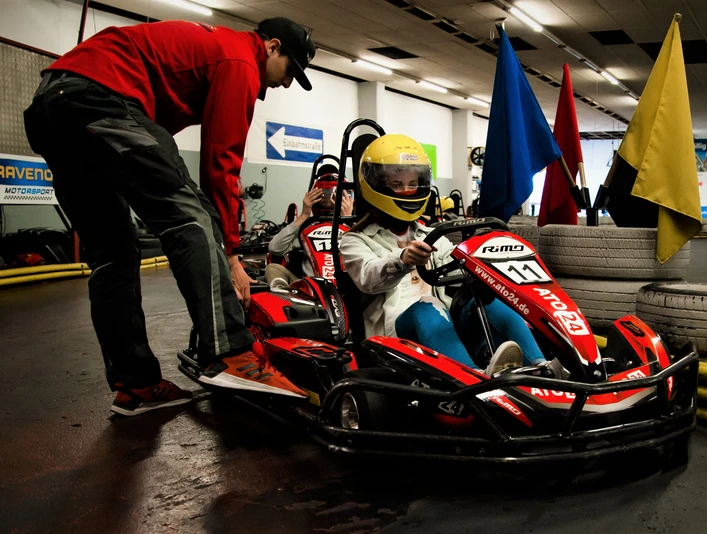 Ein Kind im gelben Helm sitzt in einem roten Go-Kart, betreut von einem Erwachsenen in einer Halle.