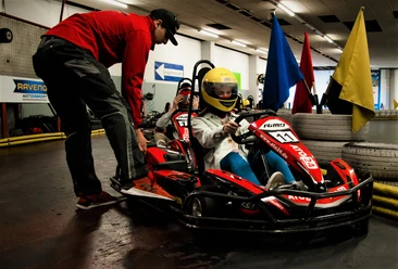 Kartbahn Werther Ein Kind im gelben Helm sitzt in einem roten Go-Kart, betreut von einem Erwachsenen in einer Halle.