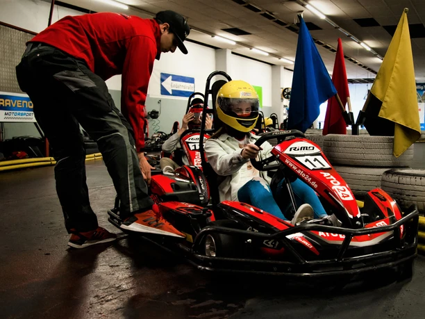Kartbahn Werther Ein Kind im gelben Helm sitzt in einem roten Go-Kart, betreut von einem Erwachsenen in einer Halle.