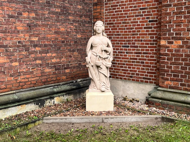 Skulptur der Justitia aus Stein mit Schwert und Waage vor einer roten Backsteinmauer.