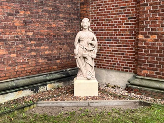 Skulptur "Justitia" Skulptur der Justitia aus Stein mit Schwert und Waage vor einer roten Backsteinmauer.