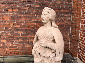 Eine steinerne Justitia-Skulptur mit Waage und Schwert, vor einer roten Backsteinmauer stehend.