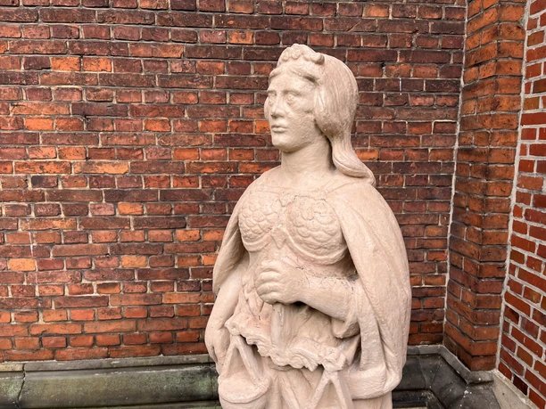 Eine steinerne Justitia-Skulptur mit Waage und Schwert, vor einer roten Backsteinmauer stehend.