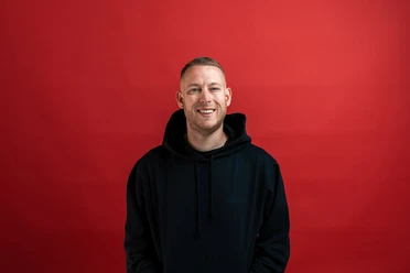 Ein junger Mann in schwarzem Hoodie vor rotem Hintergrund lächelt freundlich in die Kamera.