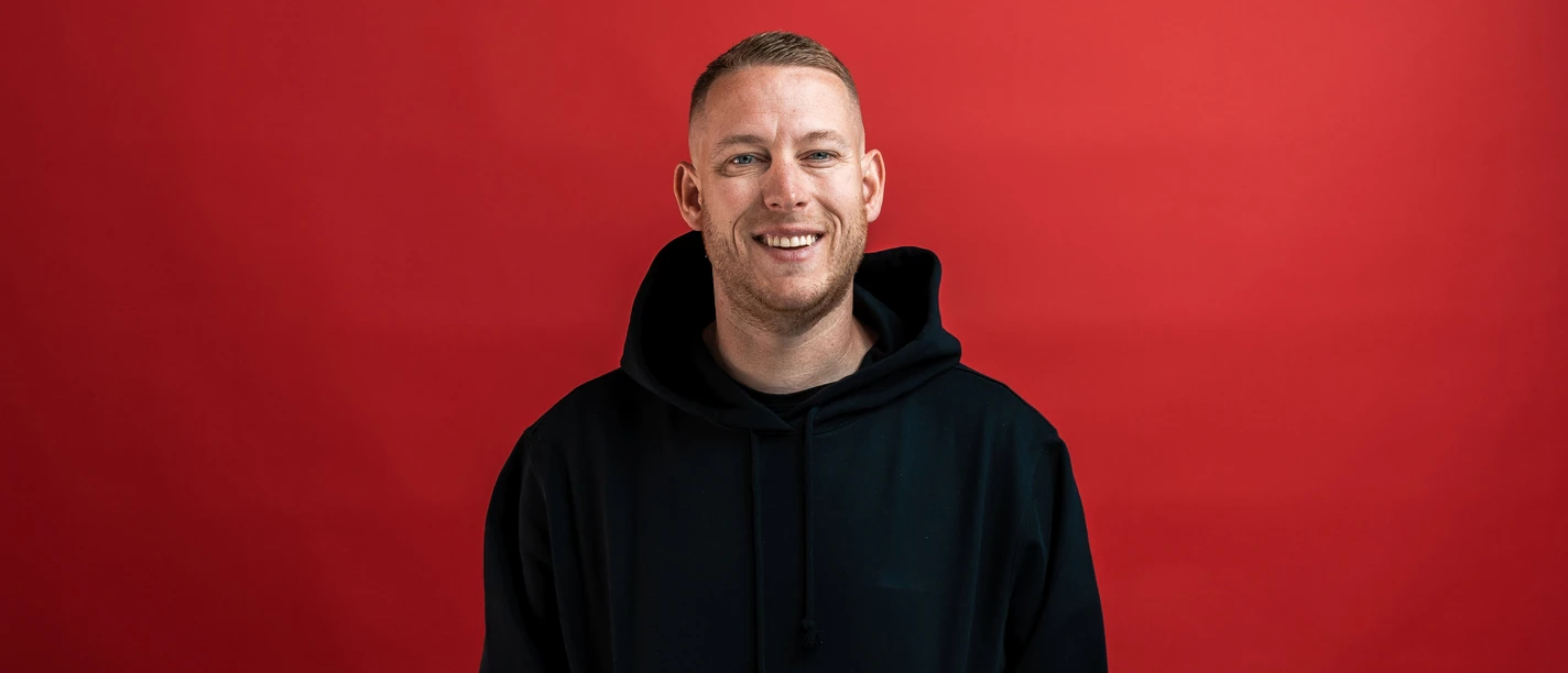 Ein junger Mann in schwarzem Hoodie vor rotem Hintergrund lächelt freundlich in die Kamera.