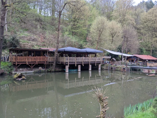 Wooden terrace with black parasols over a pond, surrounded by trees and hills, peaceful atmosphere.