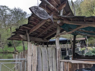 Houten hut met bordje "Forellenräucherei" in een rustieke omgeving, omgeven door bomen en een vijver. De sfeer is rustig en landelijk.
