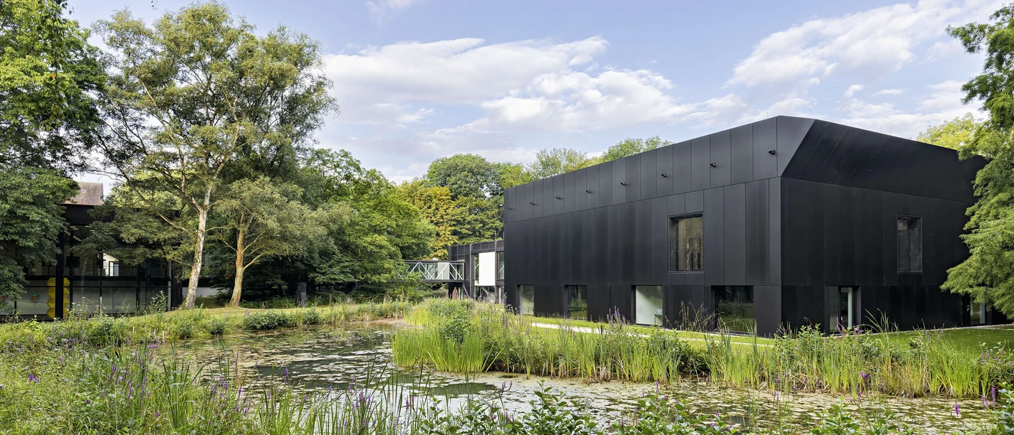 Modernes schwarzes Museumsgebäude am Teich, umgeben von Grünflächen und hohen Bäumen.