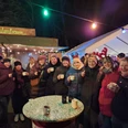Kleiner Weihnachtsmarkt.jpeg Menschen genießen warme Getränke auf einem festlichen Weihnachtsmarkt mit bunten Lichtern.