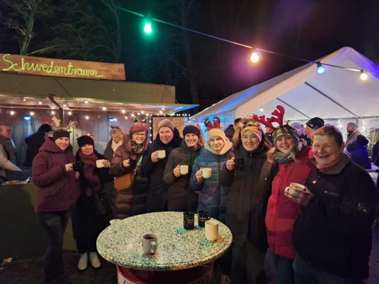 Kleiner Weihnachtsmarkt.jpeg Menschen genießen warme Getränke auf einem festlichen Weihnachtsmarkt mit bunten Lichtern.