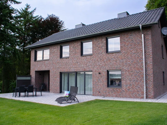 Ferienhaus Blankenrode Ziegelhaus mit Terrasse, Liegestuhl und Esstisch auf gepflegtem Rasen, umgeben von hohen Bäumen.