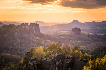 Sonnenuntergang über einer bewaldeten Landschaft mit Felsen und sanften Hügeln im Hintergrund.