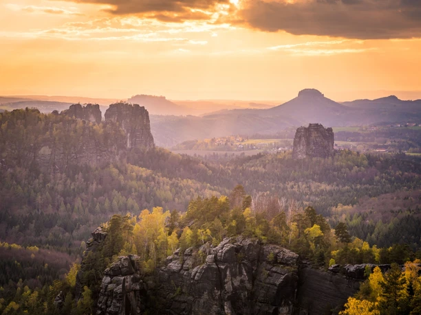 Sonnenuntergang über einer bewaldeten Landschaft mit Felsen und sanften Hügeln im Hintergrund.