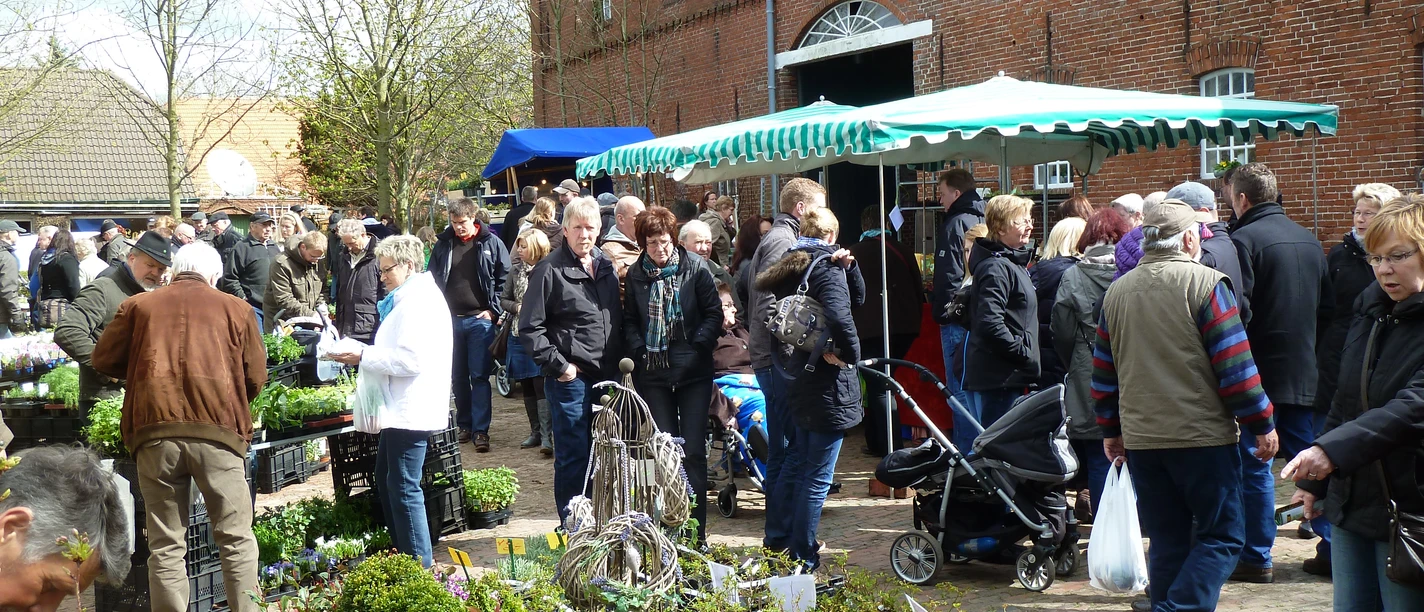 Gartentagimpressionen Besucher schlendern über einen Pflanzenmarkt vor einem Backsteinhaus mit Ständen und Frühjahrsblüten.