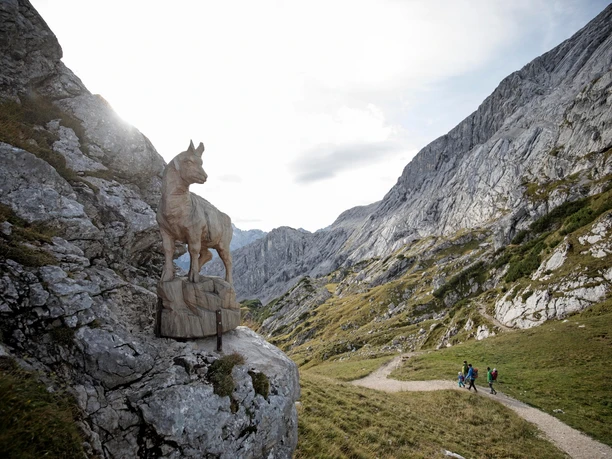 In der Felswelt der Alpspitze Holzskulptur eines Wildtiers an Felswand über einem alpinen Weg mit wandernder Gruppe
