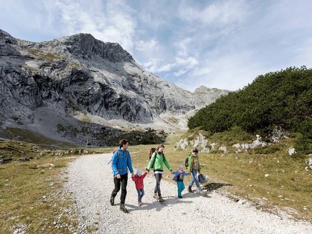 Genuss Weg Familiengruppe wandert auf breitem Schotterweg vor steilen Felswänden und lichten Bergwiesen