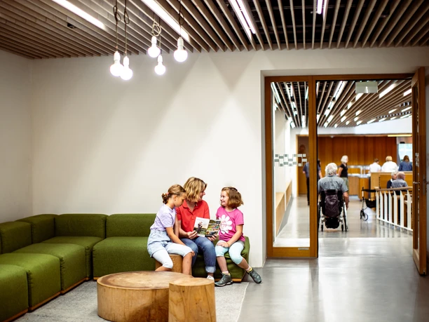 Familie im Haus Altenberg <p>Mutter mit zwei Kindern liest in einer modernen Bibliothek mit grüner Couch und Holztisch.</p>