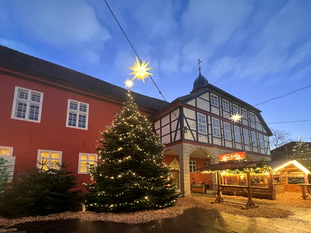 Adventszauber Nienburg (3).jpg Historic half-timbered building with festively illuminated Christmas tree and Christmas market stall.