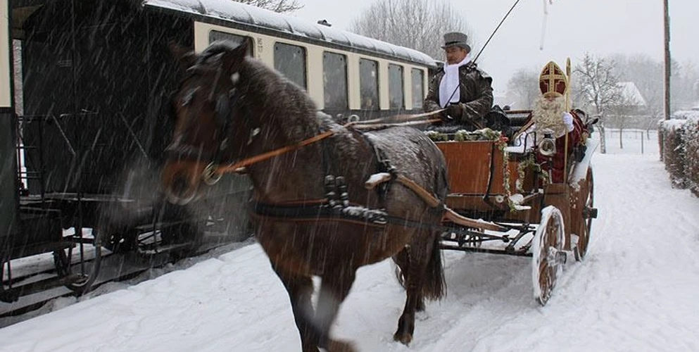 Eine historische Dampflokomotive zieht einen offenen Waggon, geschmückt mit einem Weihnachtsmann und einem Mann in traditioneller Kleidung, durch eine verschneite Winterlandschaft.