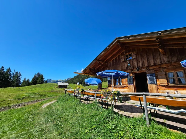 Enningalm Holzhütte mit Sitzbänken und blauen Schirmen vor grüner Almwiese und Bergkulisse im Sonnenschein