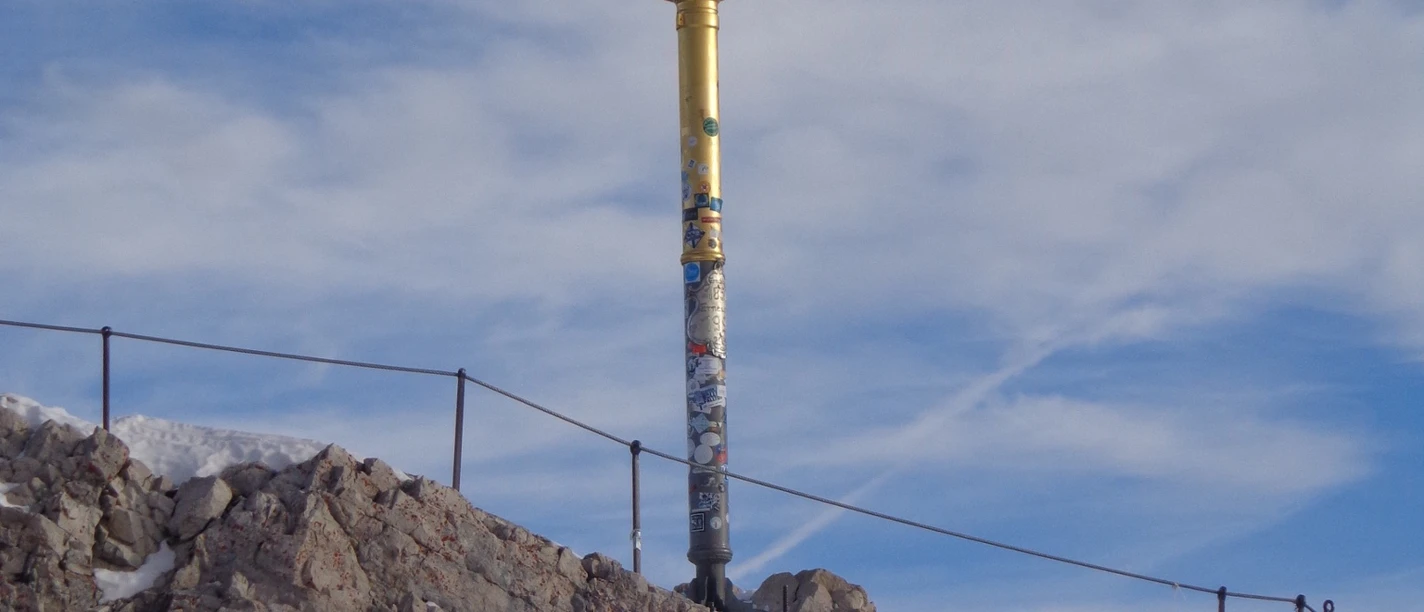 Zugspitzkreuz Du siehst ein goldenes Kreuz auf einem Fels mit Schnee.