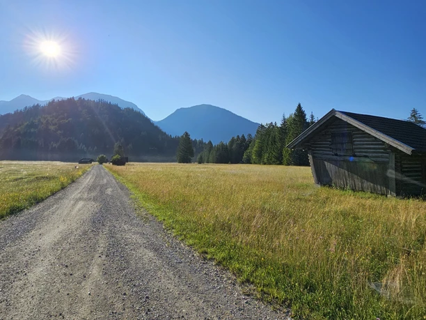 Reschbergwiesen Schotterweg durch weite Wiese mit Holzhütte, im Hintergrund bewaldete Berge unter Morgensonne