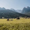 Familie Radl Familie beim Radfahren auf Wiesenweg vor Alpenkulisse mit verstreuten Hütten.