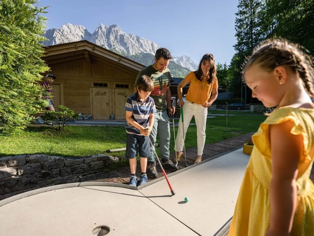 Familie spielt Minigolf vor Holzhütte und Bergen bei sonnigem Wetter