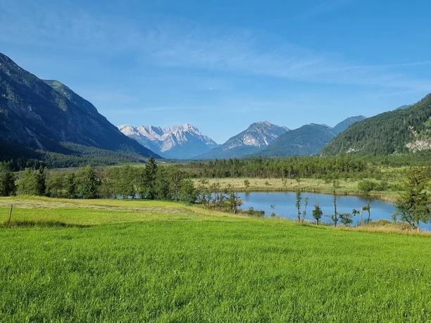 Blick von Garmisch-Partenkirchen ins Loisachtal Weite Wiese vor kleinem See mit bewaldeten Hängen und hellen Berggipfeln unter klarem Himmel