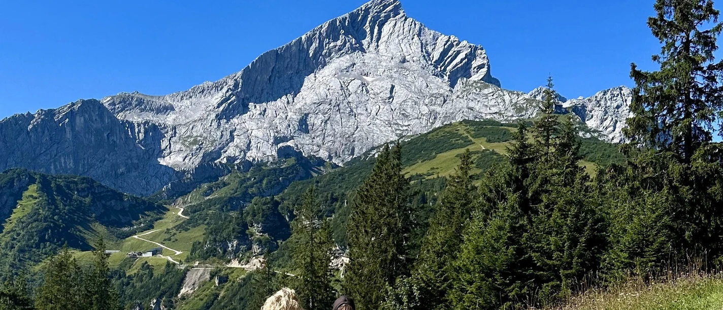 Am Kreuzeck Zwei Wandernde auf breitem Weg vor markanter Felskulisse unter blauem Himmel.