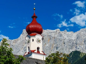 Ehrwald Kirchturm mit roter Zwiebelhaube vor steiler Bergkulisse unter blauem Himmel.