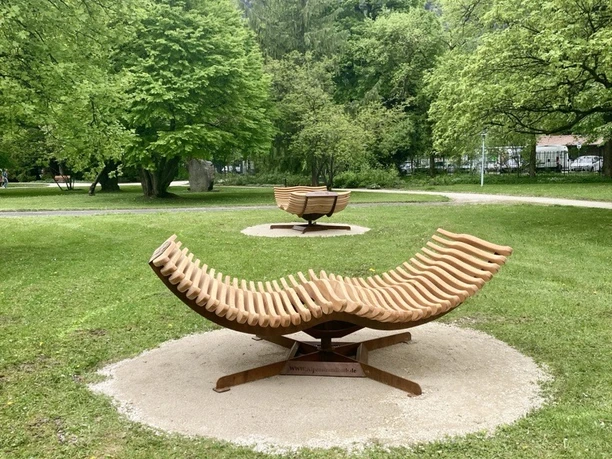Familien Sonnenliegen Geschwungene Holzliegen auf einer Wiese vor dichtem Grün und fernen Berggipfeln