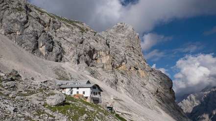Berghütte unter steilen Felswänden im sonnigen alpinen Gelände mit teils wolkigem Himmel.