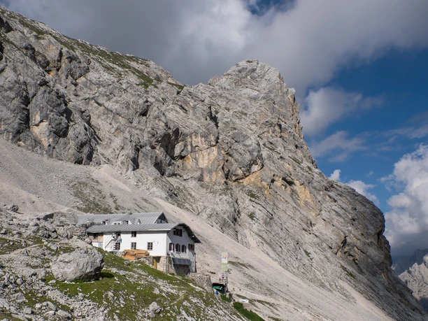 Knorrhütte Berghütte unter steilen Felswänden im sonnigen alpinen Gelände mit teils wolkigem Himmel.