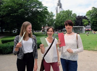 Drei Frauen in einem Park halten bunte Karten in der Hand und lächeln in die Kamera.