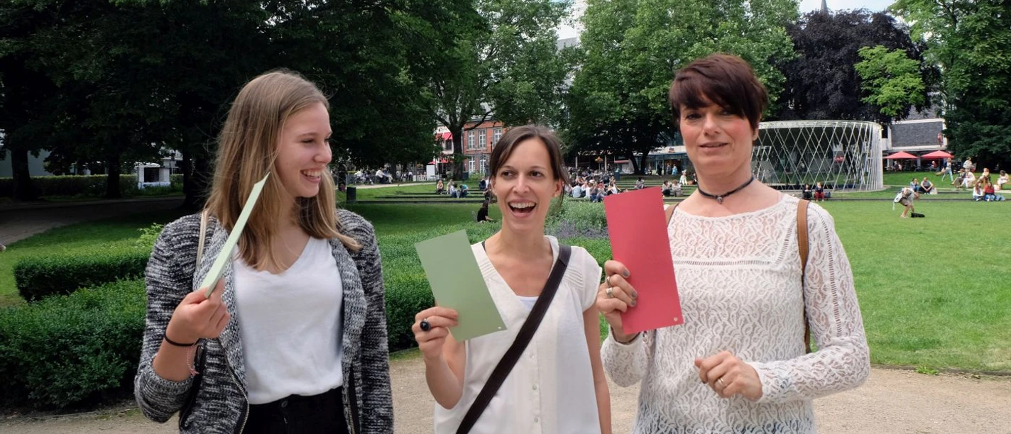 Die Lügenführung in Aachen Drei Frauen in einem Park halten bunte Karten in der Hand und lächeln in die Kamera.